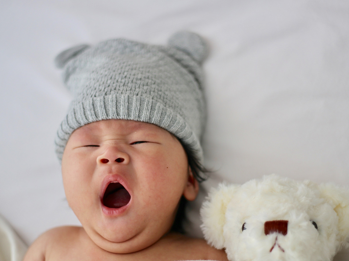 Newborn child wearing a bear hat yawning as they lay in bed under the covers with a teddy bear next to them