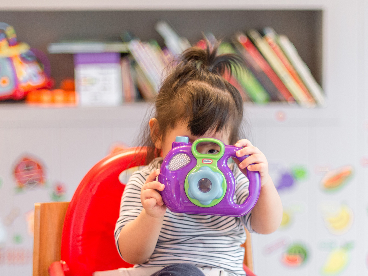 Child holding a toy camera to her face while sitting in a high chair in a childcare facility