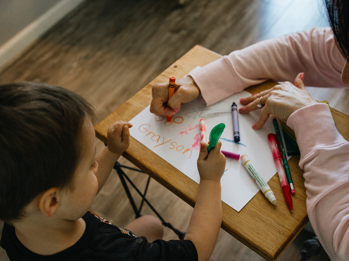 Woman helping a young boy write his name and color in crayons