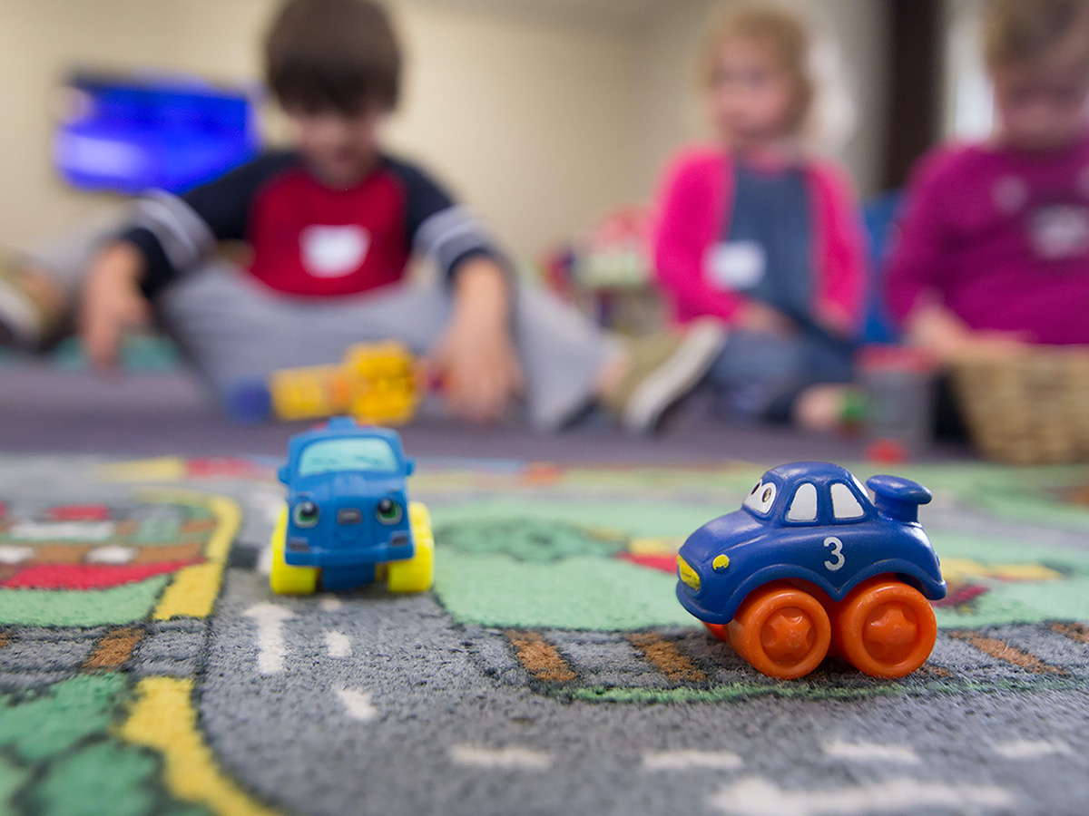 Close up of toy cars on a city map style carpet with children playing in the background
